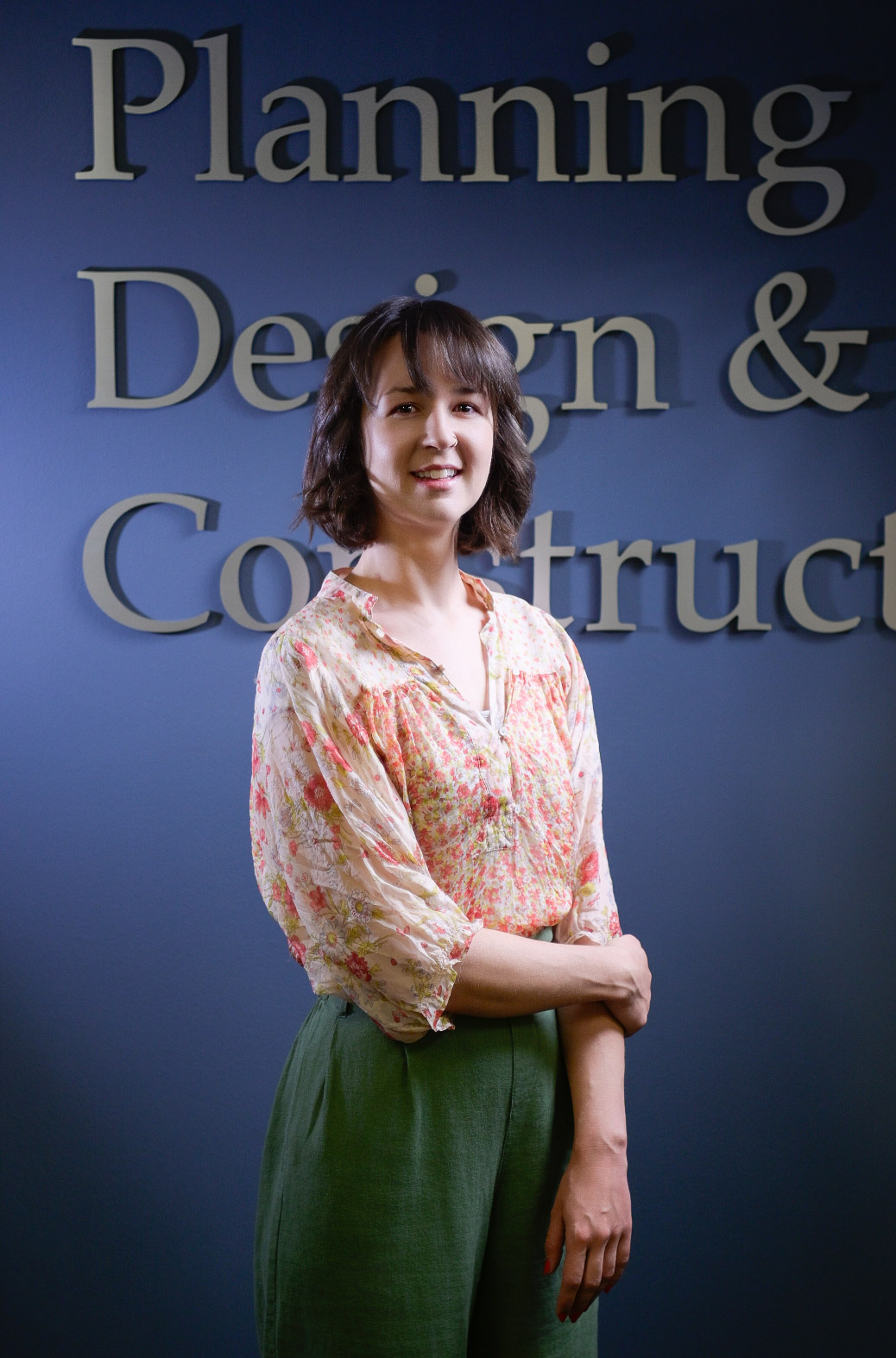 Woman with shoulder-length brown hair smiling, wearing a floral blouse and green pants, against a dark blue wall with embossed lettering.