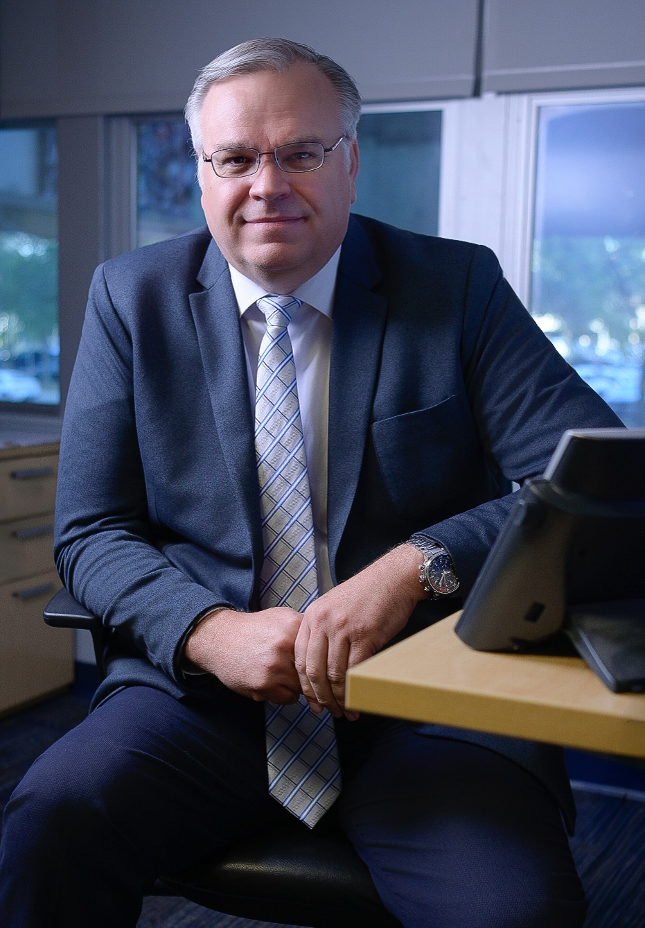 Man in a suit and tie sits facing forward, resting arms on a desk. He wears glasses and a watch. A window is visible in the background.