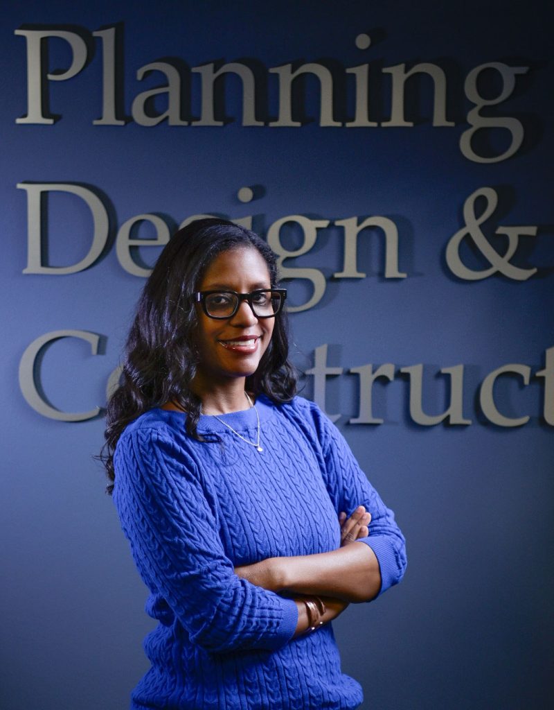 Woman with long dark curly hair wearing glasses and a blue sweater, arms crossed, smiling at the camera. Blue wall with large white text beh