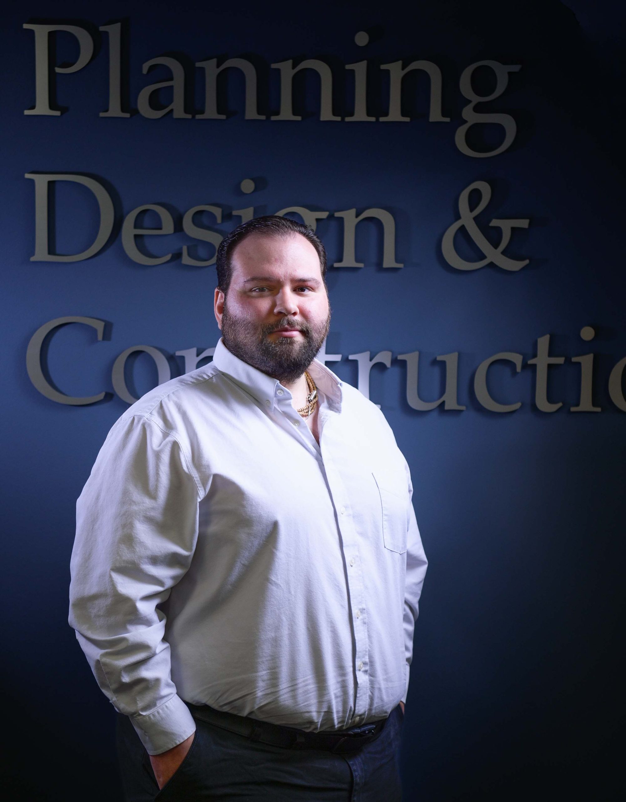 Man with beard in white shirt, hands in pockets, smiles at camera. Dark blue background with blurred, large white text.